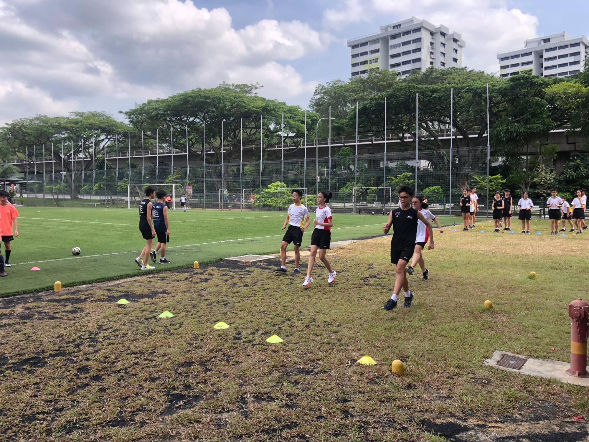 Students run and exercise on a grass field beside a fenced soccer pitch, with trees and tall buildings in the background.